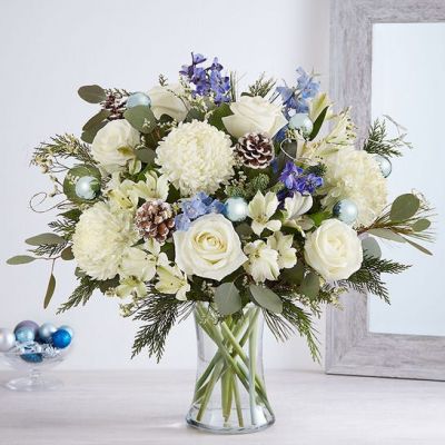 Send wonderful winters wishes with our luxurious arrangement. Snowy white blooms are gathered with lush greenery, accents of blue, gray and glistening silver & gold. Loosely gathered in a glass vase, it’s a gift designed with stunning seasonal style.